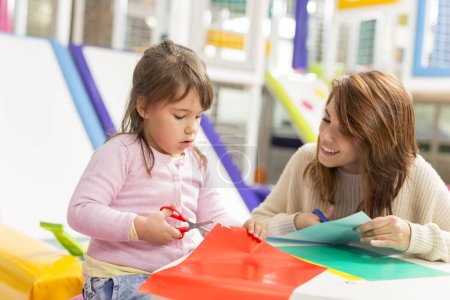 Téléchargez les photos : Mère et fille assises sur un plancher de salle de jeux, coupant du papier de collage coloré avec des ciseaux et s'amusant. Focus sur la fille - en image libre de droit