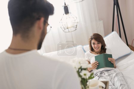 Photo for Happy and surprised young woman lying in bed in the morning while her husband is bringing her breakfast and flowers on a tray - Royalty Free Image