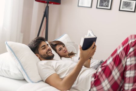 Photo for Couple in love lying in bed, hugging and reading books after waking up, enjoying the weekend morning. Focus on the girl - Royalty Free Image