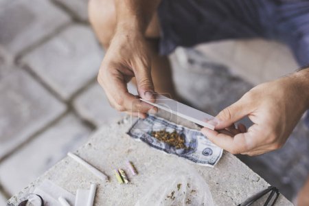 Photo for Detail of male hands rolling a joint with rolling paper and cannabis buds in the background - Royalty Free Image