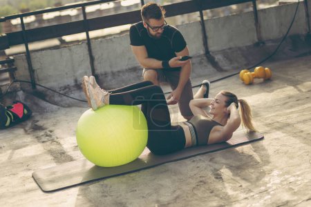 Foto de Mujer haciendo abdominales con las piernas levantadas en una pelota de pilates con la supervisión de su instructor personal de fitness - Imagen libre de derechos