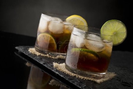 Photo for Cuba Libre cocktails with brown rum, lemon juice, coke and ice cubes, decorated with fresh lime slice on modern black stone tray - Royalty Free Image