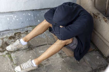 Foto de Deprimido, joven desesperado sentado en la terraza de la azotea del edificio, con capucha, sosteniendo la cabeza en las manos - Imagen libre de derechos