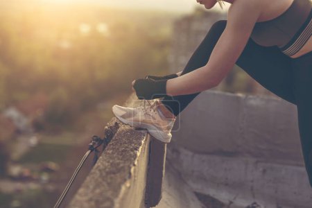 Photo for Woman in a sportswear standing on a building rooftop terrace, tying shoelaces before workout. Focus on the sneakers - Royalty Free Image