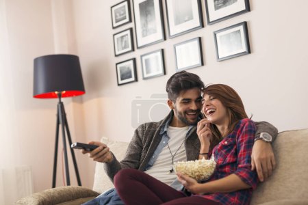 Photo for Couple in love sitting on a living room sofa, watching TV and eating popcorn, man switching channels with remote control. Focus on the man - Royalty Free Image