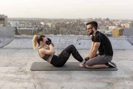Photo for Attractive woman in sportswear doing sit ups on a building rooftop terrace with the assistance of a personal fitness instructor - Royalty Free Image