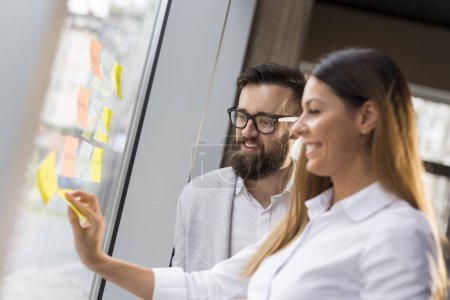 Téléchargez les photos : Homme d'affaires et femme d'affaires brainstorming en utilisant des notes collantes sur un mur de verre dans un bureau. Concentre-toi sur l'homme - en image libre de droit