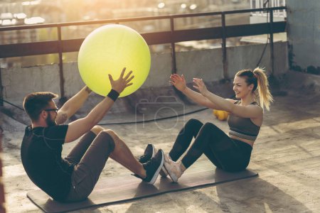Photo for Couple sitting on a yoga mat on a building rooftop terrace, doing crunches with a pilates ball. Focus on the girl - Royalty Free Image