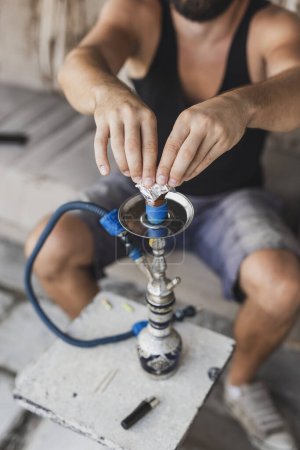 Photo for Detail of male hands placing tinfoil over a fruity flavored, molasses based hokkah tobacco, getting narghile ready for use - Royalty Free Image