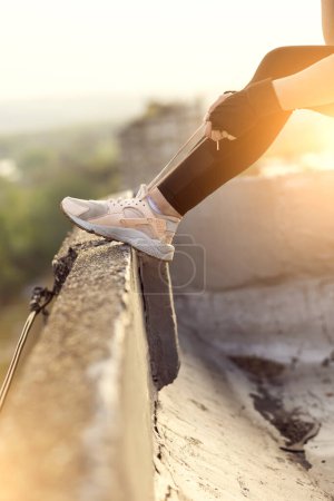 Téléchargez les photos : Femme portant des vêtements de sport debout sur un toit-terrasse du bâtiment, attachant des lacets avant l'entraînement - en image libre de droit