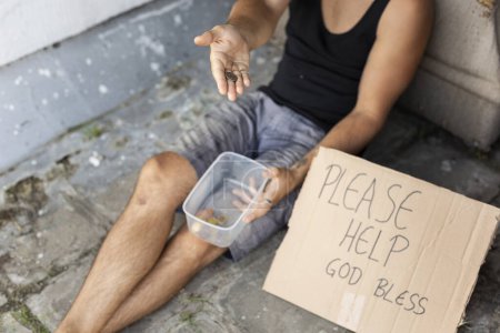 Foto de Vista de alto ángulo de un hombre sin hogar sentado en la calle, pidiendo dinero. Concéntrate en la moneda en la mano - Imagen libre de derechos