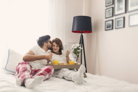 Foto de Pareja feliz enamorada acostada en la cama, desayunando y disfrutando de su tiempo juntos, chico besando a la chica - Imagen libre de derechos