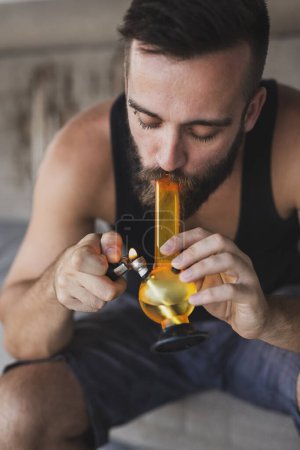 Téléchargez les photos : Jeune homme fumant de l'herbe à l'aide de bong ; homme inhalant de la vapeur de marijuana d'un bong - en image libre de droit