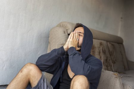 Téléchargez les photos : Jeune homme déprimé et désespéré assis sur le toit-terrasse de l'immeuble, portant un sweat à capuche, tenant la tête dans les mains - en image libre de droit