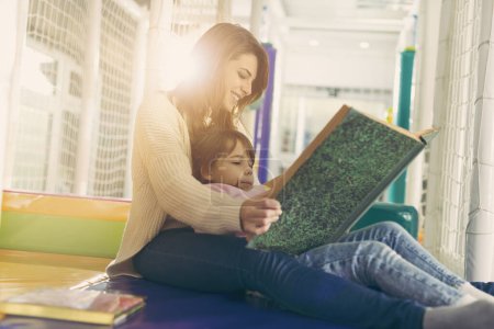 Téléchargez les photos : Mère et fille assises dans une salle de jeux, lisant un conte de fées ; fille assise sur les genoux de la mère - en image libre de droit