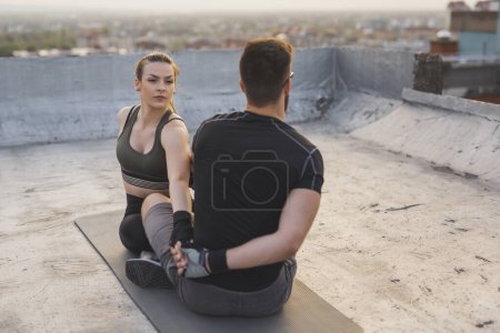 Foto de Pareja en una ropa deportiva, sentado en una estera de yoga, estirándose antes de un entrenamiento de terraza en la azotea del edificio - Imagen libre de derechos