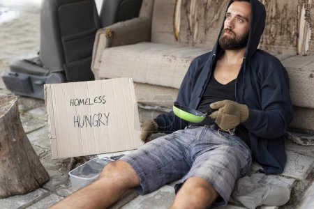 Photo for Beggar wearing hoodie over cap and fingerless gloves, sitting in the street, begging for money - Royalty Free Image