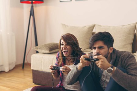 Photo for Couple in love enjoying their free time, sitting on the living room floor, playing video games and having fun. Focus on the woman - Royalty Free Image