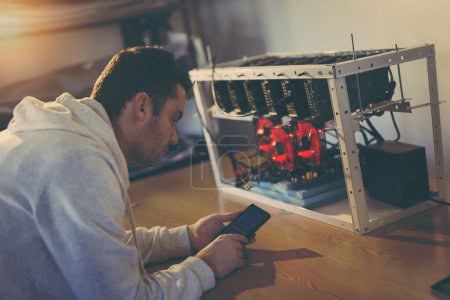 Photo for Programmer setting up a mining rig for cryptocurrency mining, holding a smart phone and adjusting the mining rates app - Royalty Free Image
