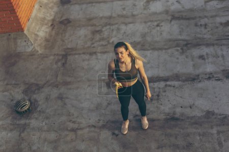 Foto de Hermosa mujer joven de pie en una terraza de la azotea del edificio, tomando un descanso de entrenamiento y comiendo plátano - Imagen libre de derechos