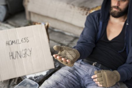Foto de Un vagabundo con capucha sobre gorra y guantes sin dedos, sentado en la calle, rogando dinero. Enfoque selectivo en la mano - Imagen libre de derechos