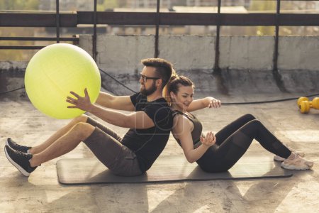 Photo for Couple sitting back to back on a yoga mat on a building rooftop terrace, exercising by passing the pilates ball to each other - Royalty Free Image