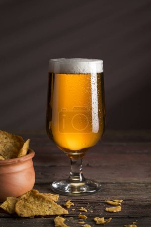 Foto de Vaso de cerveza fría pálida con un tazón de tortillas fritas sobre una mesa rústica de madera - Imagen libre de derechos