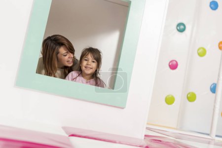 Photo for Mother and daughter playing in a playroom, hiding in a small wooden house, whispering and storytelling. Focus on the daughter - Royalty Free Image