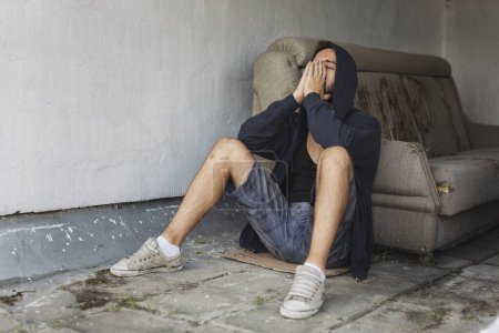 Téléchargez les photos : Jeune homme déprimé et désespéré assis sur le toit-terrasse de l'immeuble, portant un sweat à capuche, tenant la tête dans les mains - en image libre de droit
