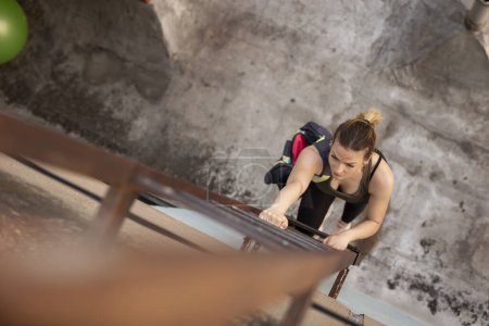 Téléchargez les photos : Femme séduisante portant des vêtements de sport et portant un sac de gymnastique grimpant une échelle d'évacuation d'incendie, allant pour un entraînement sur le toit d'un bâtiment extérieur - en image libre de droit