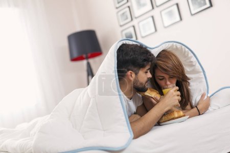 Foto de Pareja enamorada acostada debajo de las sábanas, tomando croissants frescos y zumo de naranja para desayunar en la cama y disfrutando del fin de semana por la mañana - Imagen libre de derechos