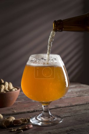 Téléchargez les photos : Un verre de bière froide non filtrée est versé et un bol d'arachides sur une table de pub rustique en bois. Concentration sélective sur la mousse - en image libre de droit