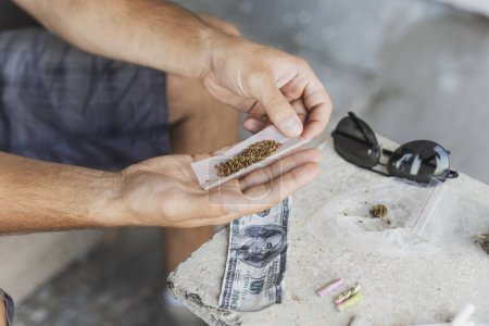 Foto de Detalle de las manos masculinas enrollando una articulación con papel de liar y brotes de cannabis en el fondo - Imagen libre de derechos