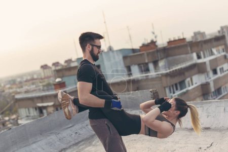 Foto de Pareja usando ropa deportiva haciendo ejercicios de crujidos colgantes en una terraza en la azotea de un edificio - Imagen libre de derechos