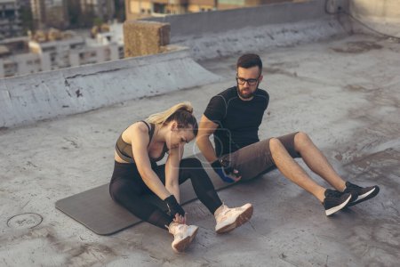 Téléchargez les photos : Couple échauffement pour un bâtiment sur le toit-terrasse séance d'entraînement, assis sur un tapis de yoga, étirement. Focus sur la femme - en image libre de droit