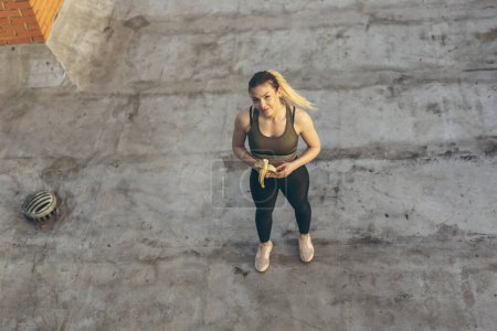 Photo for High angle view of a beautiful young woman standing on a building rooftop terrace, taking a workout break and eating banana - Royalty Free Image