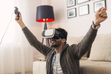Téléchargez les photos : Jeune homme assis sur le sol du salon, portant des lunettes VR et jouant à un jeu VR - en image libre de droit