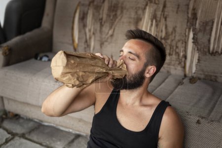 Photo for Drunkard sitting on a building rooftop terrace, holding a bottle in a paper bag, drinking - Royalty Free Image