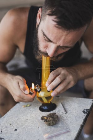 Téléchargez les photos : Jeune homme fumant de l'herbe à l'aide de bong ; homme inhalant de la vapeur de marijuana d'un bong - en image libre de droit