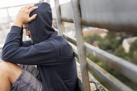 Photo for Depressed, desperate young man sitting on the building rooftop terrace, wearing hoodie, holding head in hands - Royalty Free Image