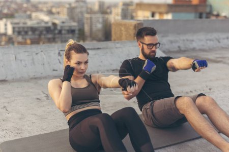 Photo for Couple wearing sportswear doing punch crunches exercise on a building rooftop terrace. Focus on the woman - Royalty Free Image