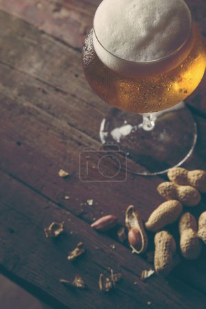 Foto de Vaso de cerveza fría pálida y algunos cacahuetes sobre una mesa rústica de madera. Enfoque selectivo en la espuma - Imagen libre de derechos