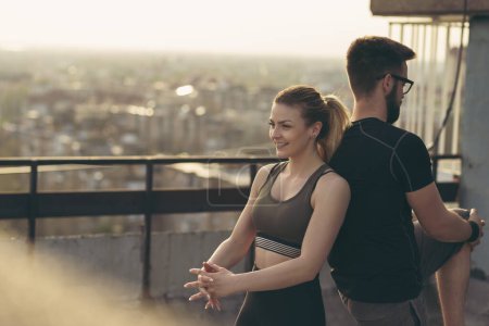 Photo for Young couple on a building rooftop terrace stretching before workout; urban skyline in the background. - Royalty Free Image
