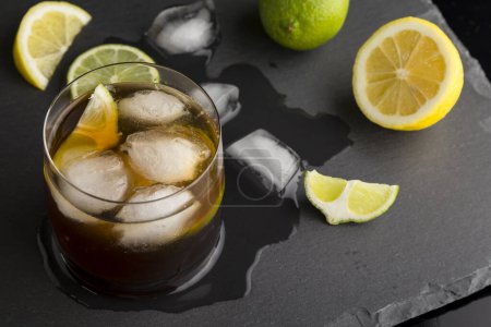 Téléchargez les photos : Vue en angle élevé d'un verre de Cuba Libre cocktail avec rhum, coke, jus de citron et glaçons sur un plateau moderne en pierre noire. Concentration sélective sur la glace - en image libre de droit