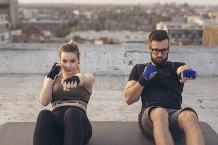 Photo for Couple wearing sportswear, sitting on a yoga mat and doing punch crunches exercise on a building rooftop terrace. Focus on the woman - Royalty Free Image