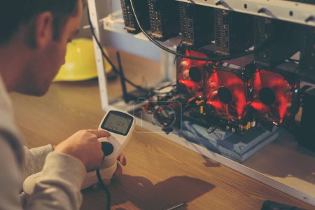 Photo for Programmer holding a wattmeter measuring cryptocurrency mining rig electrical energy consumption. Focus on the man's finger - Royalty Free Image