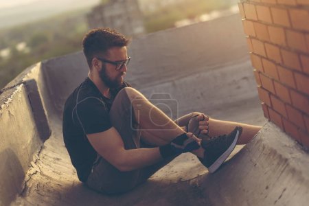 Photo for Man in a sportswear sitting on a building rooftop terrace, tying shoelaces before workout - Royalty Free Image
