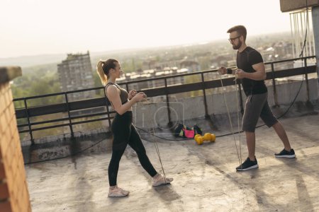 Foto de Pareja de pie en una terraza de la azotea del edificio, haciendo ejercicio con cuerdas. skyline urbano en el fondo; centrarse en la chica - Imagen libre de derechos