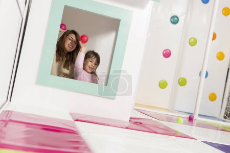 Photo for Mother and daughter playing in a playroom, hiding in a small wooden house, throwing balls through the window - Royalty Free Image