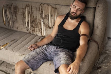 Foto de Se desmayó drogadicto sentado en el sofá después de inyectarse heroína; drogadicto con sobredosis por inyección intravenosa de heroína - Imagen libre de derechos